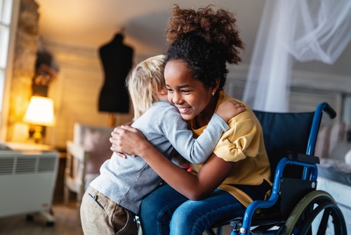 An inclusive girl in a wheelchair is hugging a boy in a living room.