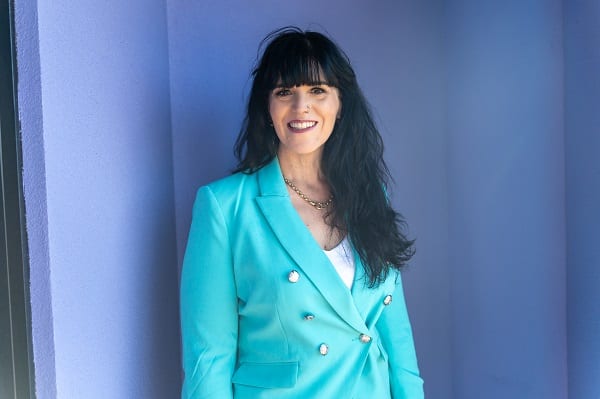 A woman in a turquoise blazer, leaning against a wall.