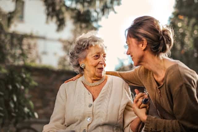 In a park, a woman is chatting with an older woman to discuss Mother's Day gift ideas.