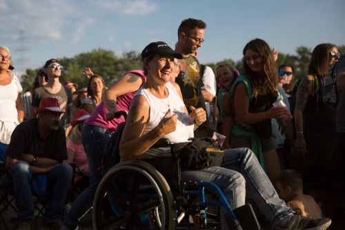 A woman in a wheelchair expressing excitement at an outdoor event surrounded by standing spectators, showing off her companion card application.