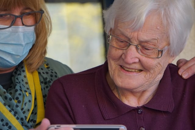 An elderly woman smiles joyfully while looking at a smartphone screen, accompanied by a masked aged care worker, likely sharing a moment of connection.