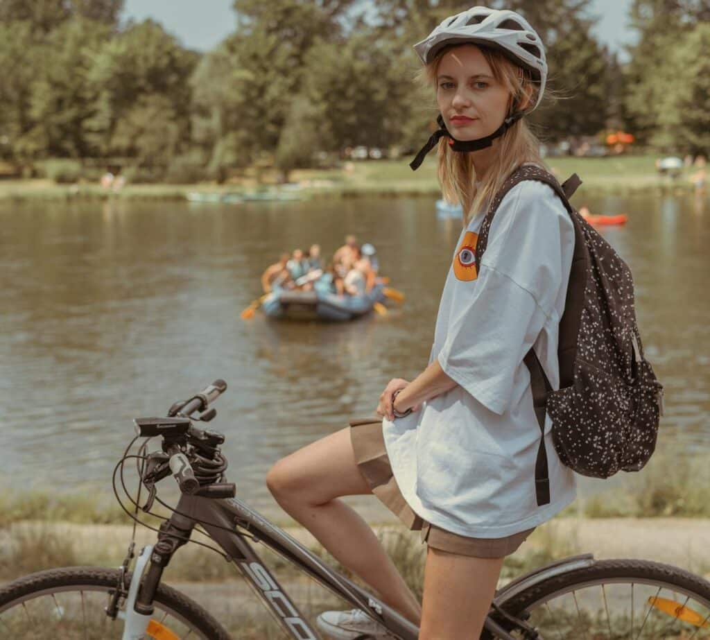 A person wearing a helmet and backpack sits on a bicycle in front of a lake with boats and people in the background, enjoying the scenery despite dealing with the symptoms of Cystic Fibrosis.