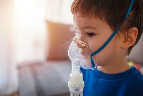 A young child in a blue shirt uses a nebulizer mask for respiratory treatment, highlighting the importance of managing cystic fibrosis.
