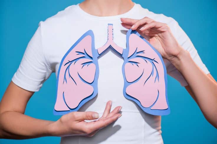 A person holds a paper cutout model of human lungs against their chest, symbolizing awareness of cystic fibrosis, with a blue background.