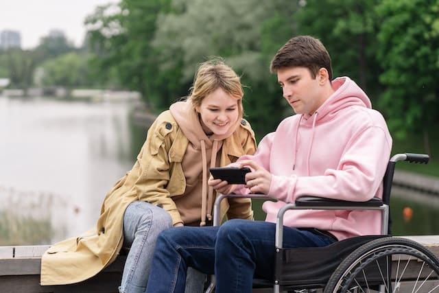 A man in a wheelchair wearing a pink hoodie shows a picture of a dog being sold via mail order online to a friend of his, a woman wearing a raincoat