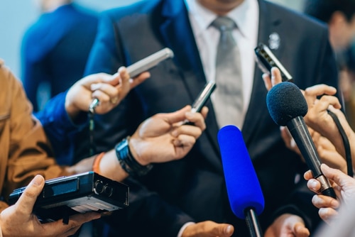 Close-up image of a person in a suit surrounded by multiple microphones and audio recording devices held by several hands, as they address the media about the NDIS scandal.