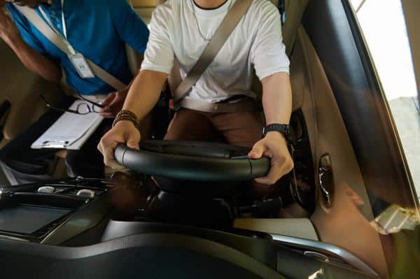 A person sits in the car, gripping the steering wheel, while another beside them holds a notebook for an OT driving assessment.