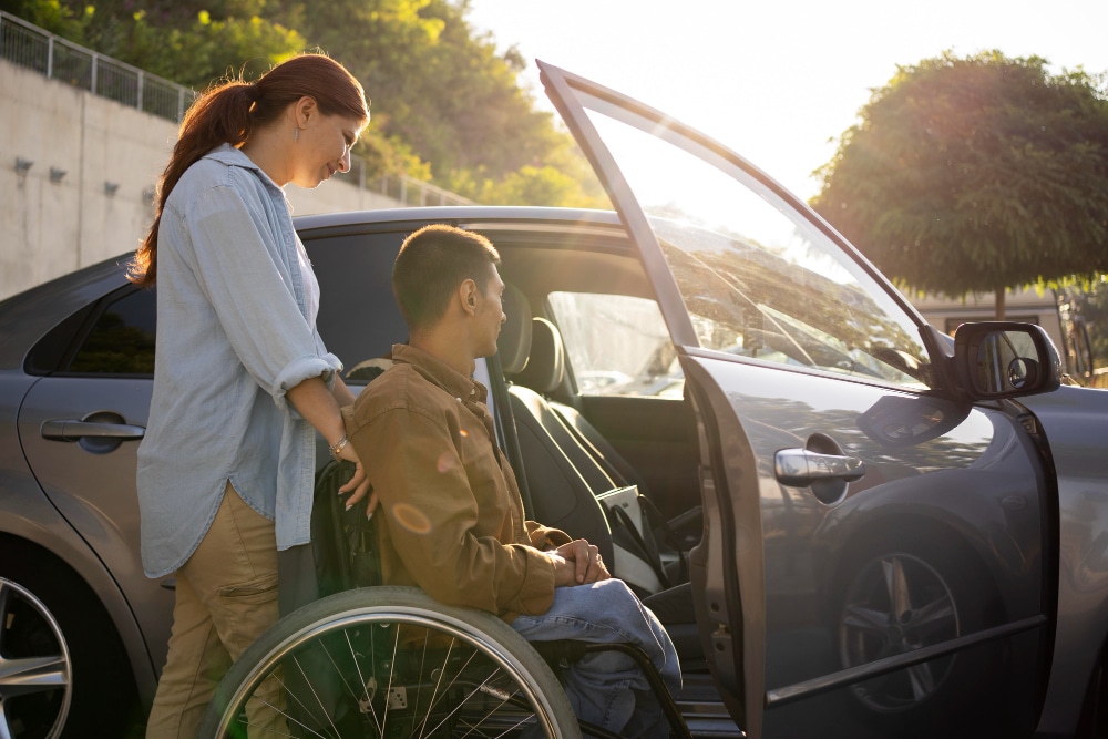 A woman, possibly a driver-trained occupational therapist, assists a man in a wheelchair as he prepares to get into a parked car with the driver's side door open on a sunny day, helping him get back on the road.