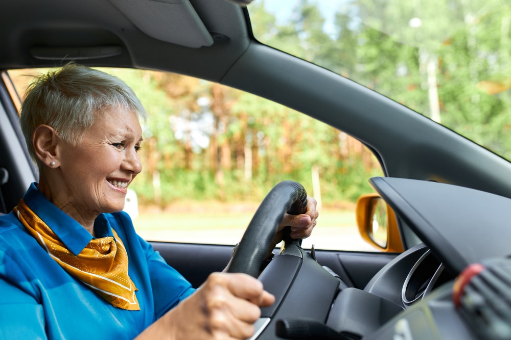 Older woman with short gray hair, wearing a blue top and yellow scarf, smiling while driving a car. As an occupational therapy success story, she enjoys her renewed freedom with trees visible through the window in the background.
