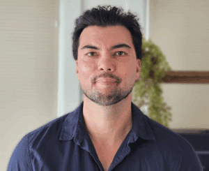 A man with short dark hair and a trimmed beard, dressed in a navy blue shirt, stands indoors with a plant and white wall in the background—he is a driver trained OT specializing in driving with disability.