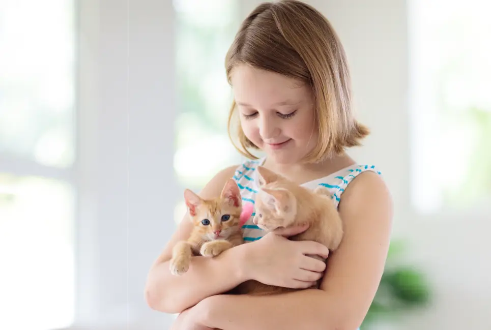 A young girl in a sleeveless dress holds two small orange kittens indoors, smiling and looking down at them—celebrating Pet Wellness during World Pet Month.